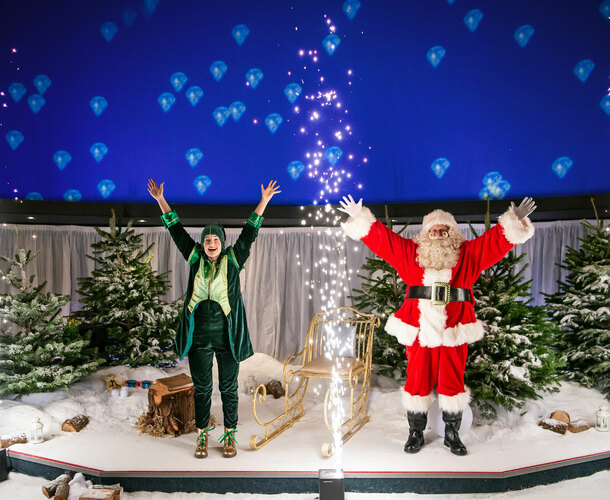 An elf and Santa stand on a stage with a sparkler going off in front of them