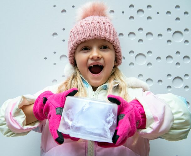 A surprised looking little girl with pink hat and gloves holds a big ice block