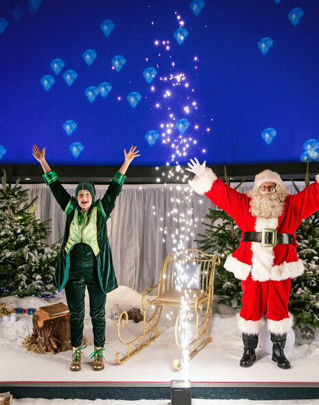 An elf and Santa stand on a stage with a sparkler going off in front of them